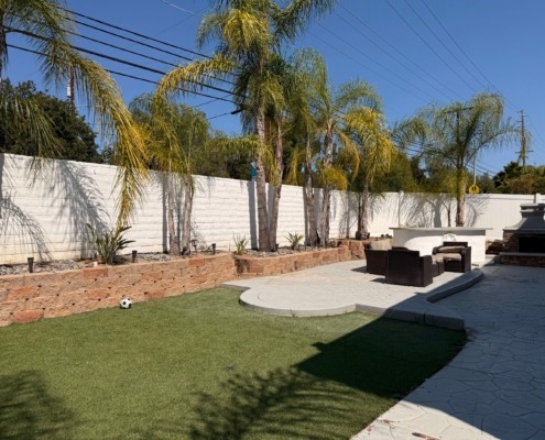 Backyard with artificial grass, palm trees, stone retaining wall, outdoor seating area, and a small soccer ball on the lawn under a clear blue sky.