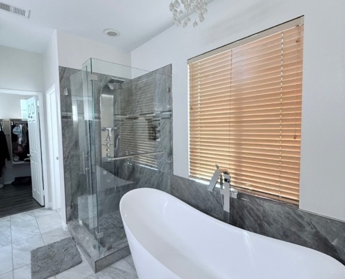 Modern bathroom with a freestanding white bathtub, glass-enclosed shower, gray tile floors, wood blinds on the window, and a small chandelier on the ceiling.